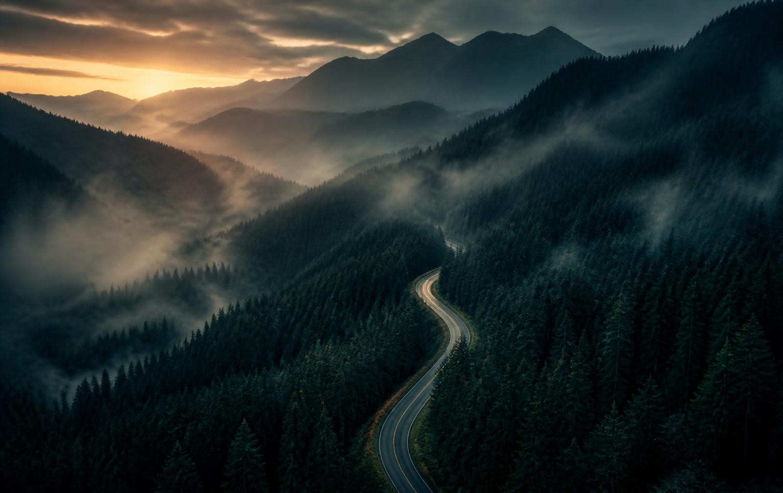 Aerial view of a road winding through misty evergreen mountains at dusk